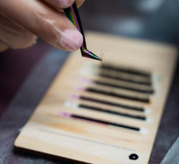 Gloved technician lifting a small lash cluster with tweezers, showing the handcrafted process