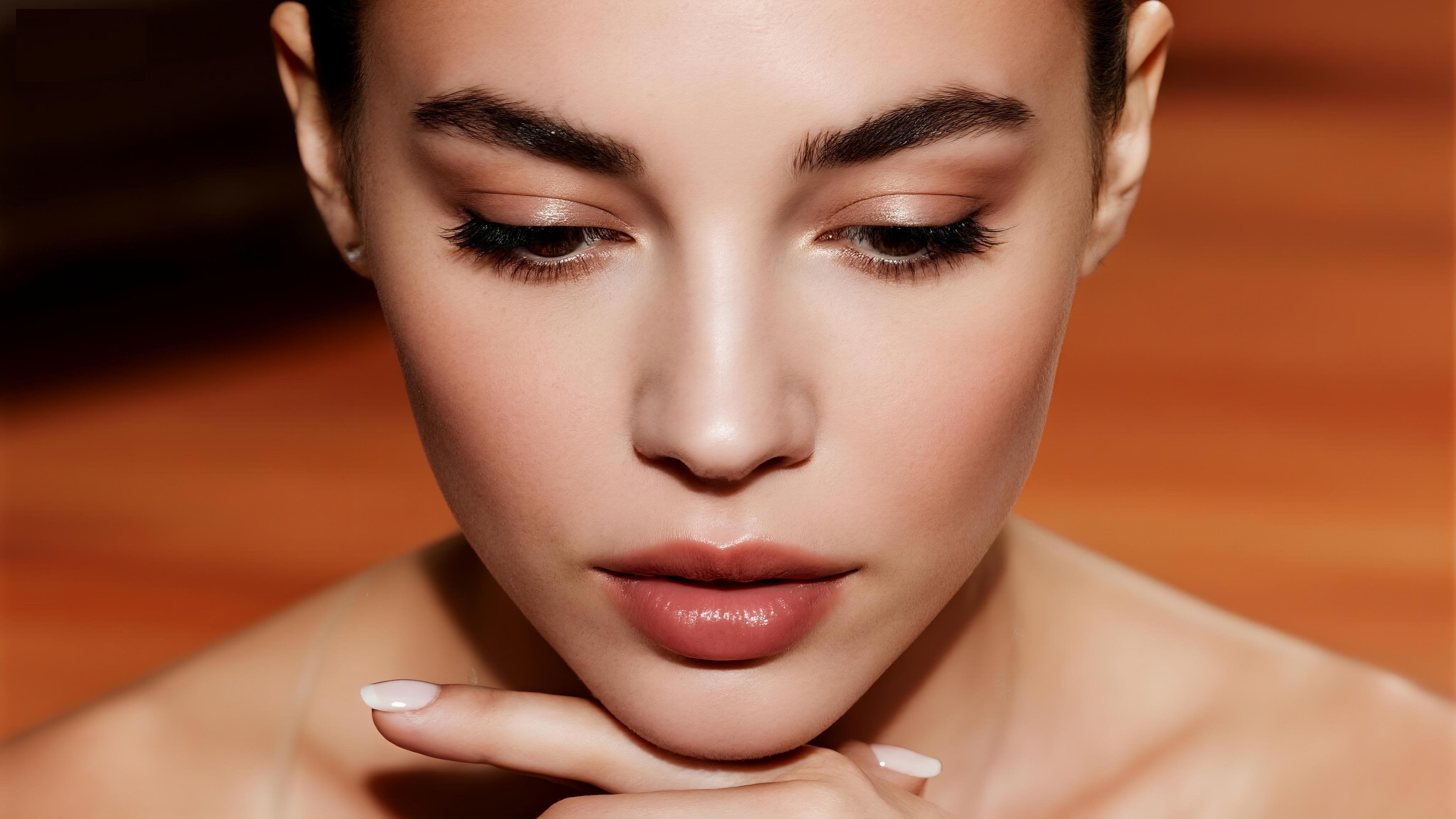 Portrait close-up with false lashes; model gazes downward with chin resting on fingertips, soft lighting and nude lips against a warm orange backdrop