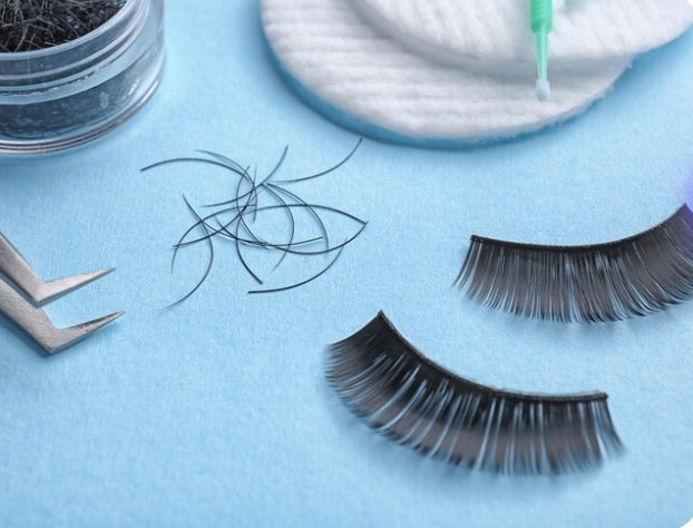 Flat lay of lash materials and tools on blue: loose fiber clusters, two strip lashes, tweezers/measure tool, cotton pads, and adhesive applicator